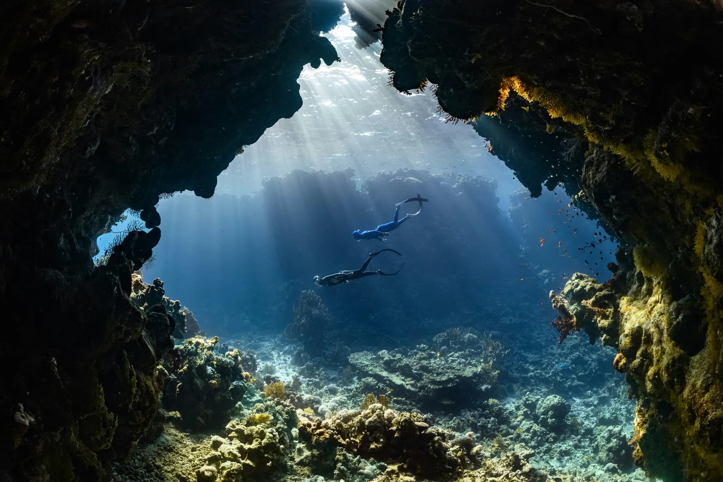 Two freedivers swimming through an underwater cave lit by sunbeams